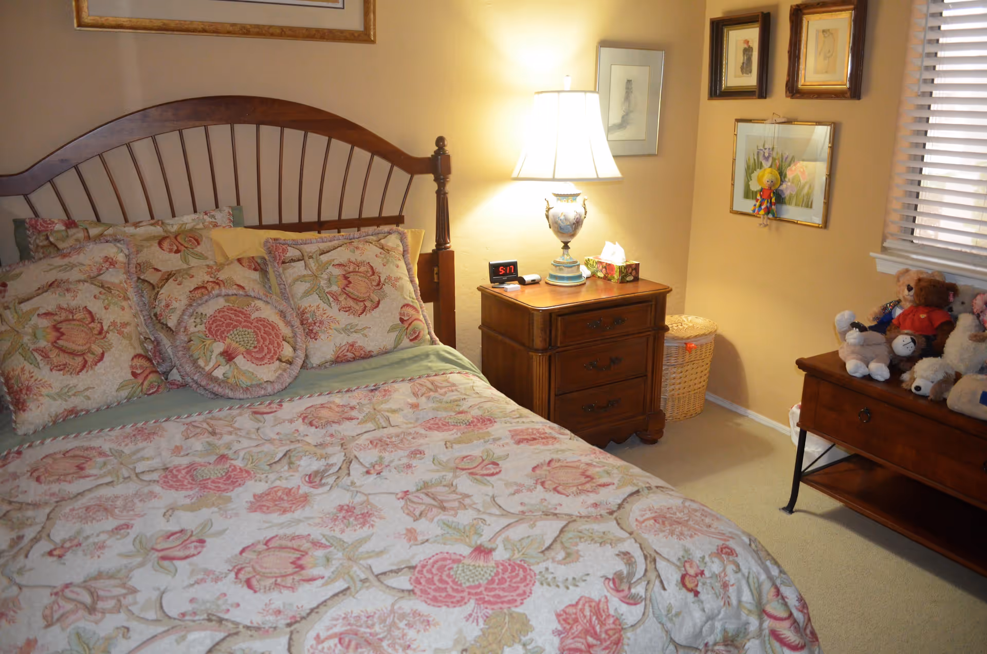 A cozy bedroom with a wooden bed frame and floral patterned bedding. There is a wooden nightstand with a decorative lamp, a digital clock, and a tissue box. The walls are adorned with framed artwork, and a window with blinds is visible. A small table holds several stuffed animals, and a wicker laundry basket is in the corner.