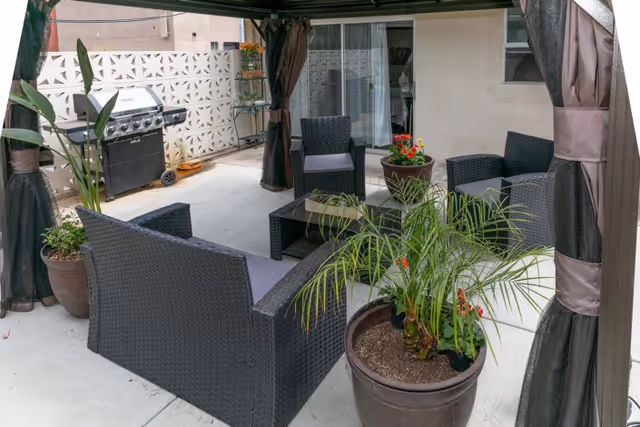 Covered patio with wicker seating, potted plants, and a gas grill outside sliding glass doors.