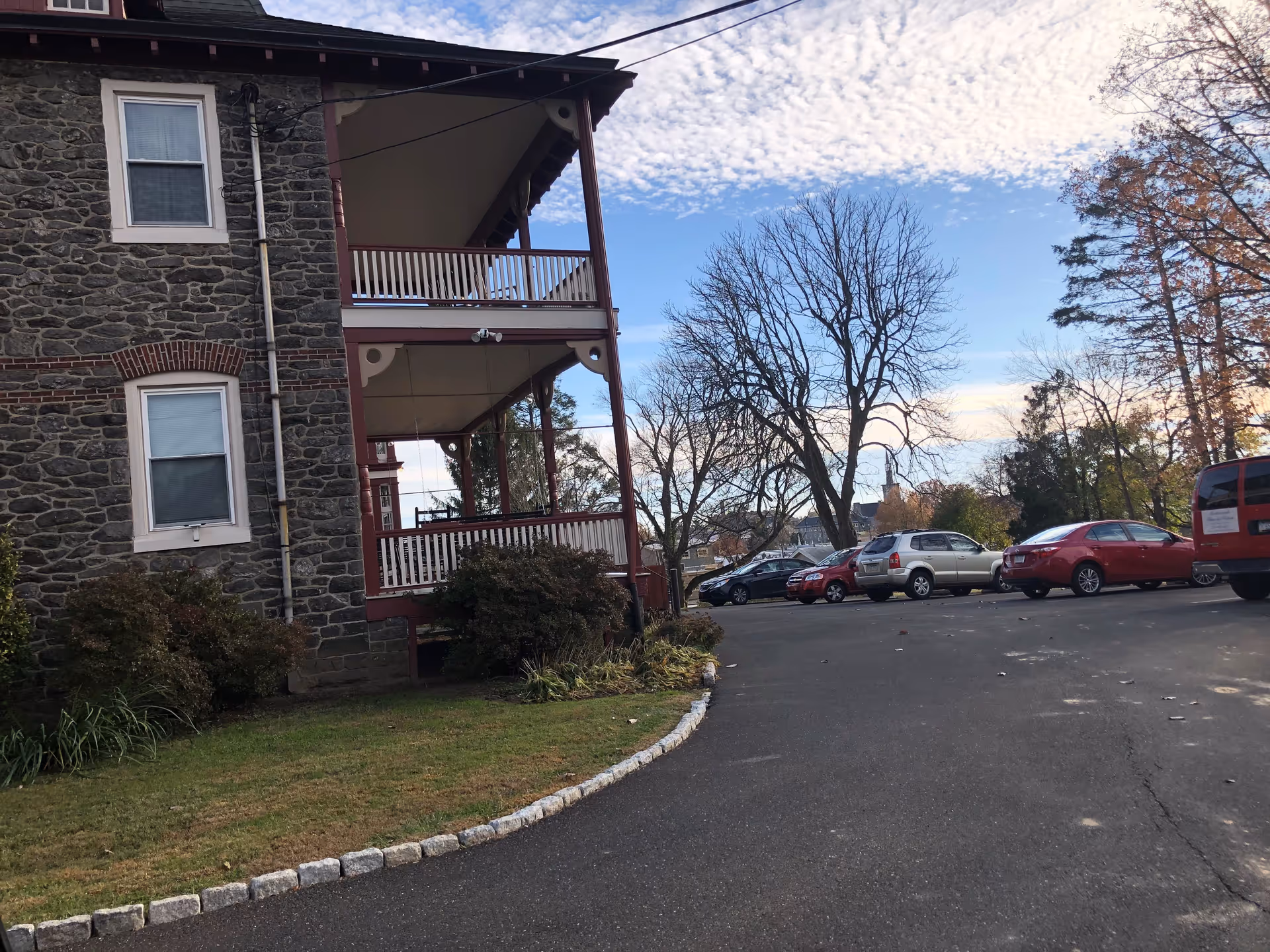 Exterior view of a stone building with two covered balconies, surrounded by a paved parking area with several parked cars and leafless trees under a partly cloudy sky.