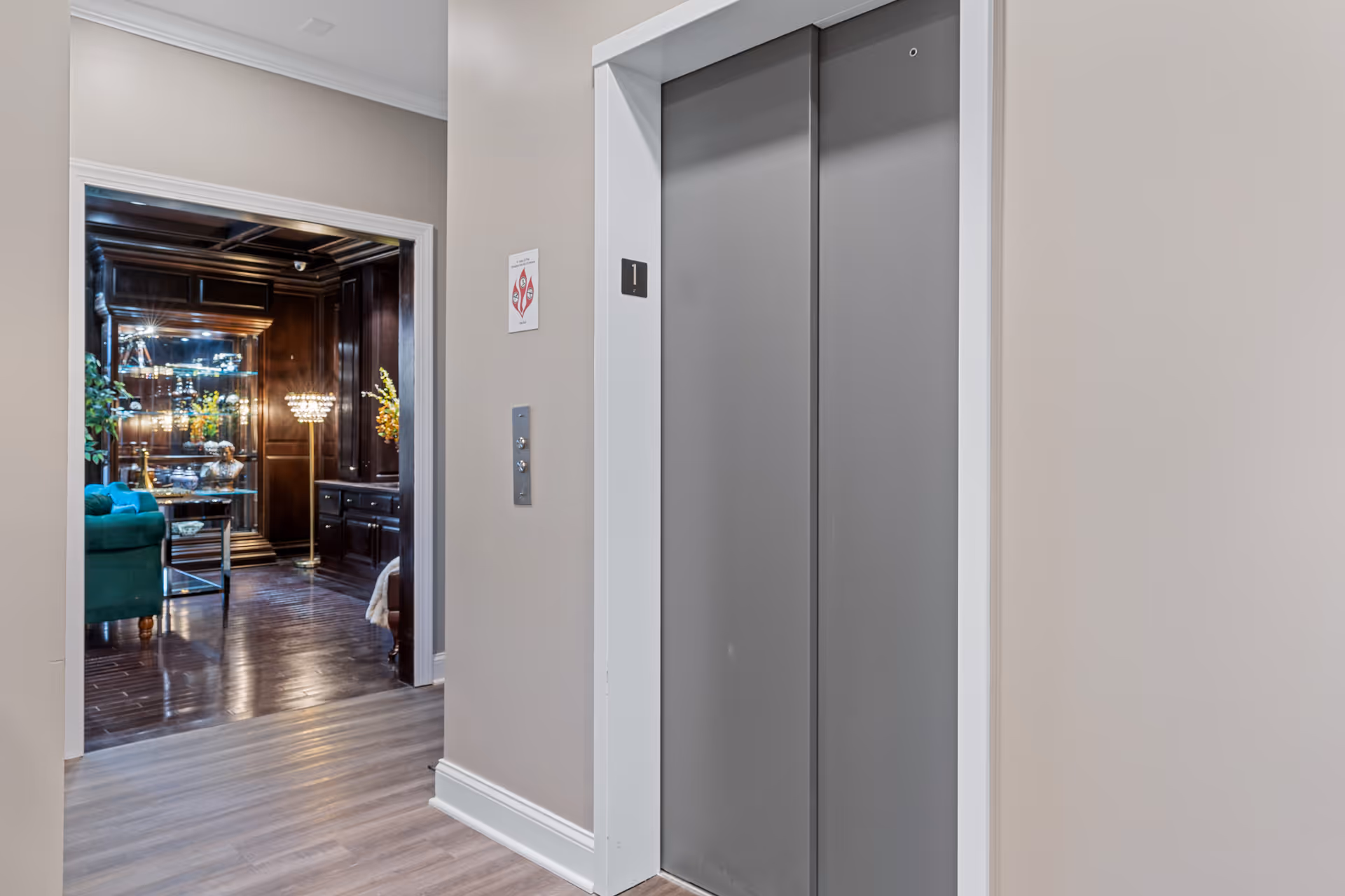 Interior view of a senior living facility hallway showing a closed elevator with gray doors and a sign indicating floor 1. To the left, there is an open doorway leading to a room with dark wood paneling, a green armchair, a glass display cabinet, and a floor lamp with a decorative shade.