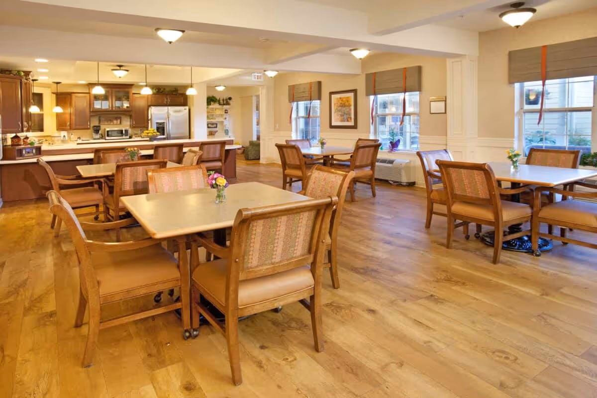 Well-lit communal dining area with multiple wooden tables and chairs and a kitchen in the background.