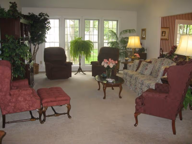 A cozy living room with large windows letting in natural light. The room features two brown recliners, a floral patterned sofa with cushions, two red armchairs with a matching footstool, a wooden coffee table with a vase of pink flowers, several potted plants, and two table lamps providing warm lighting.