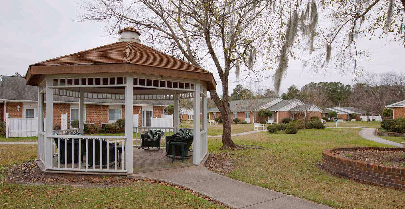 Outdoor gazebo with chairs inside situated in a grassy courtyard area surrounded by single-story brick buildings and trees with bare branches.