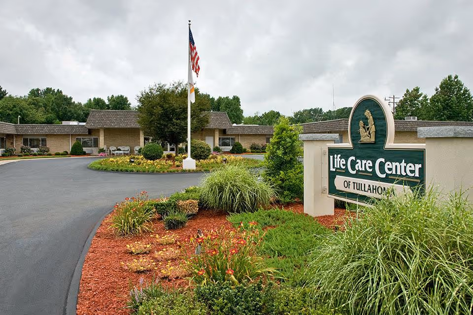Entrance of Life Care Center of Tullahoma showing the facility sign, driveway, flagpole, and landscaped flower beds.