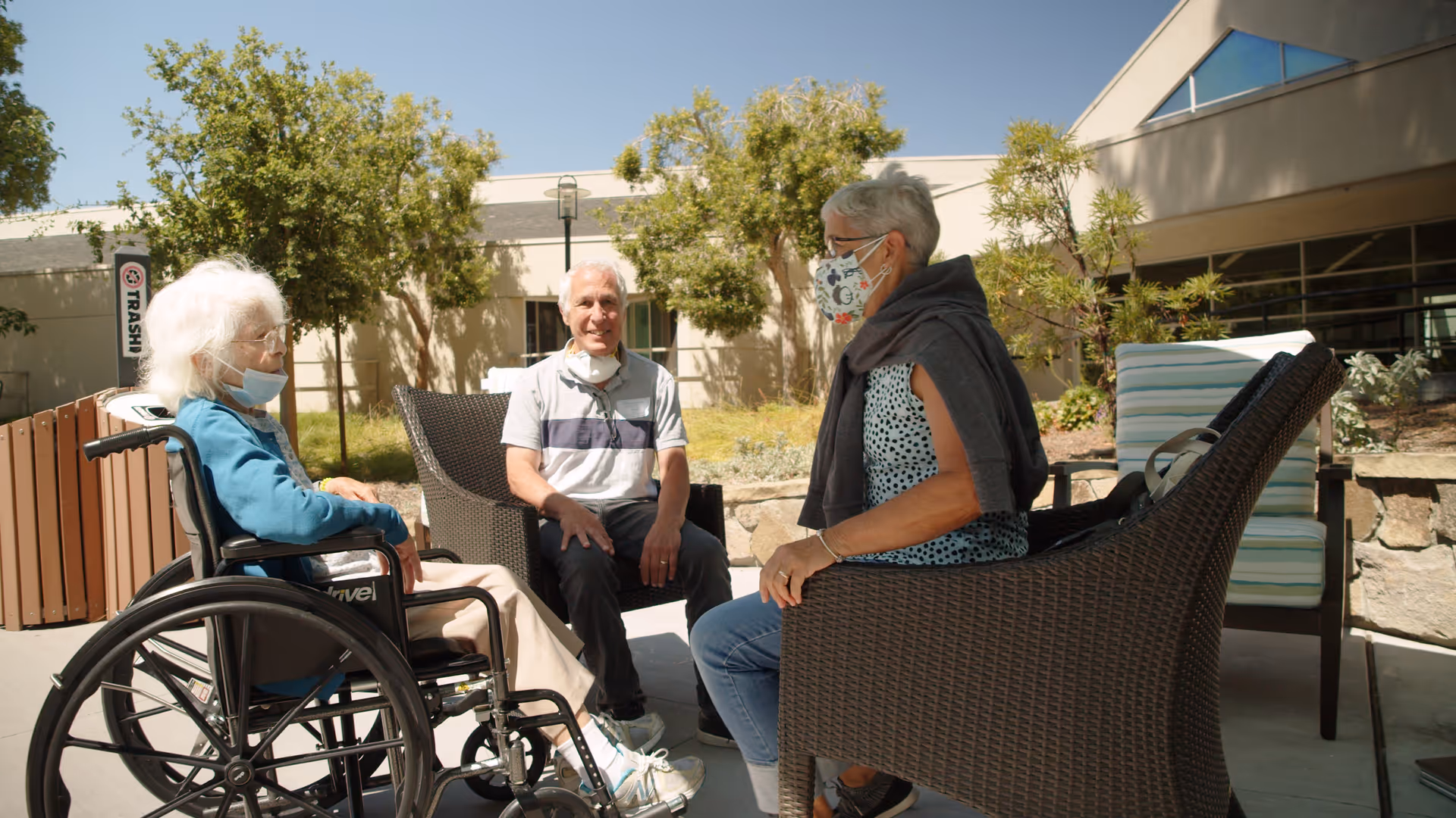 Three elderly people sitting and talking outdoors at Mercy Retirement & Care Center. One woman is in a wheelchair wearing a blue jacket and a face mask under her chin. A man in a gray and white striped shirt is sitting on a chair smiling. Another woman with short gray hair, wearing a patterned face mask and a sleeveless top with a sweater draped over her shoulders, is seated on a wicker chair. They are surrounded by trees and a building under a clear blue sky.