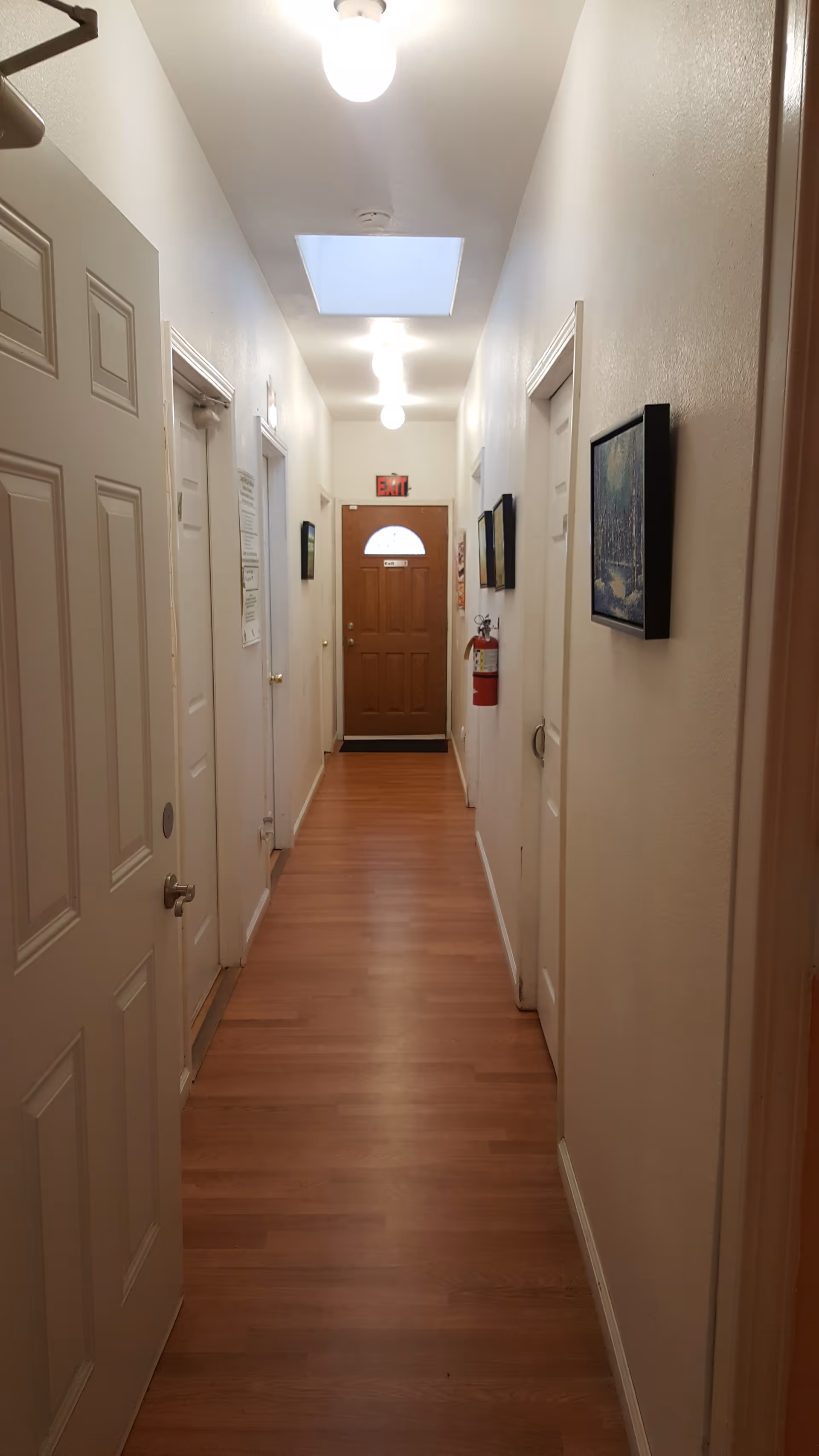 Narrow hallway with wooden floor, white walls and doors on both sides, ceiling lights, and a wooden exit door at the far end.