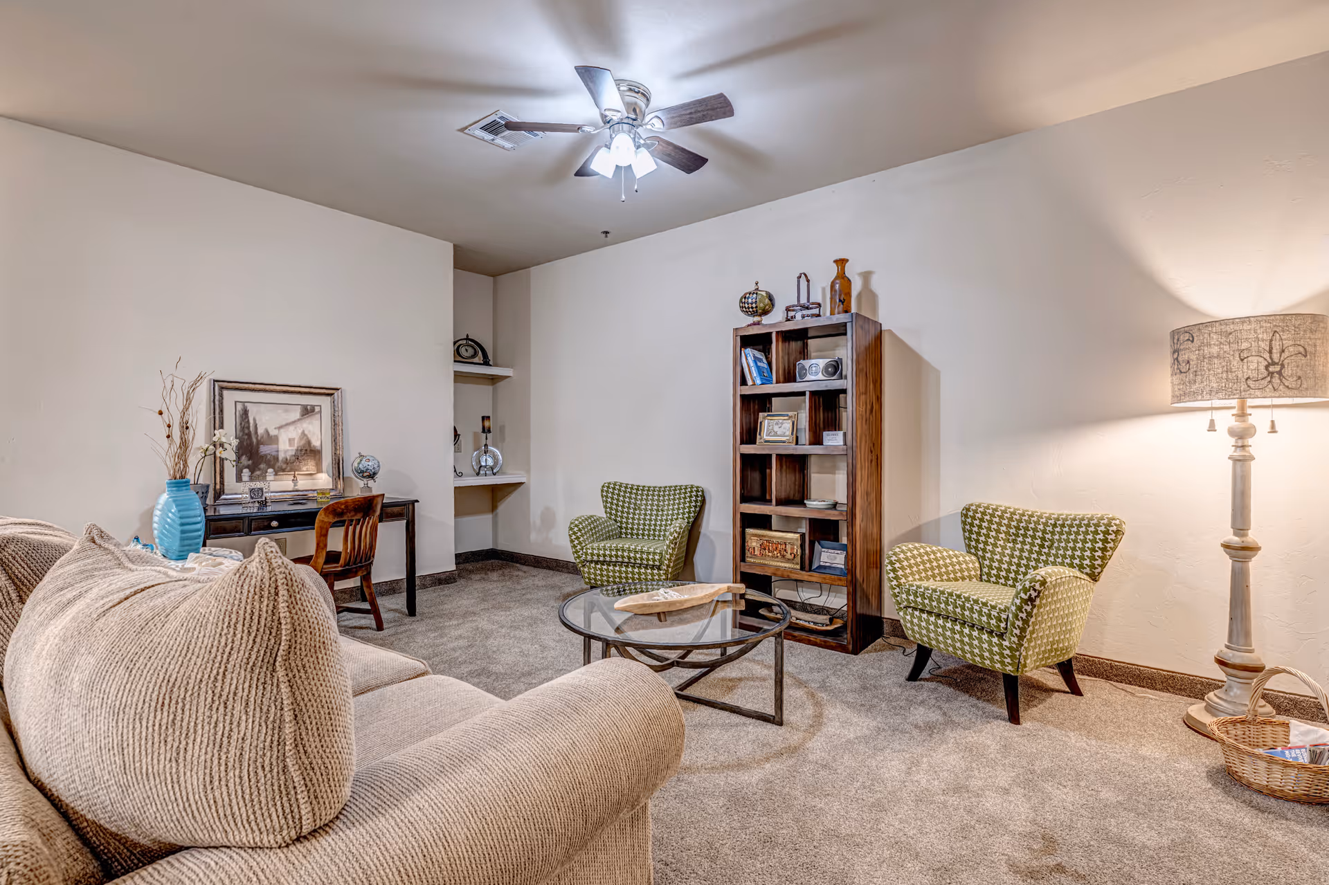 A cozy living room with a beige sofa, two green patterned armchairs, a round glass coffee table, a wooden bookshelf with decorative items, a floor lamp with a patterned shade, and a small desk with a chair and framed artwork on the wall.