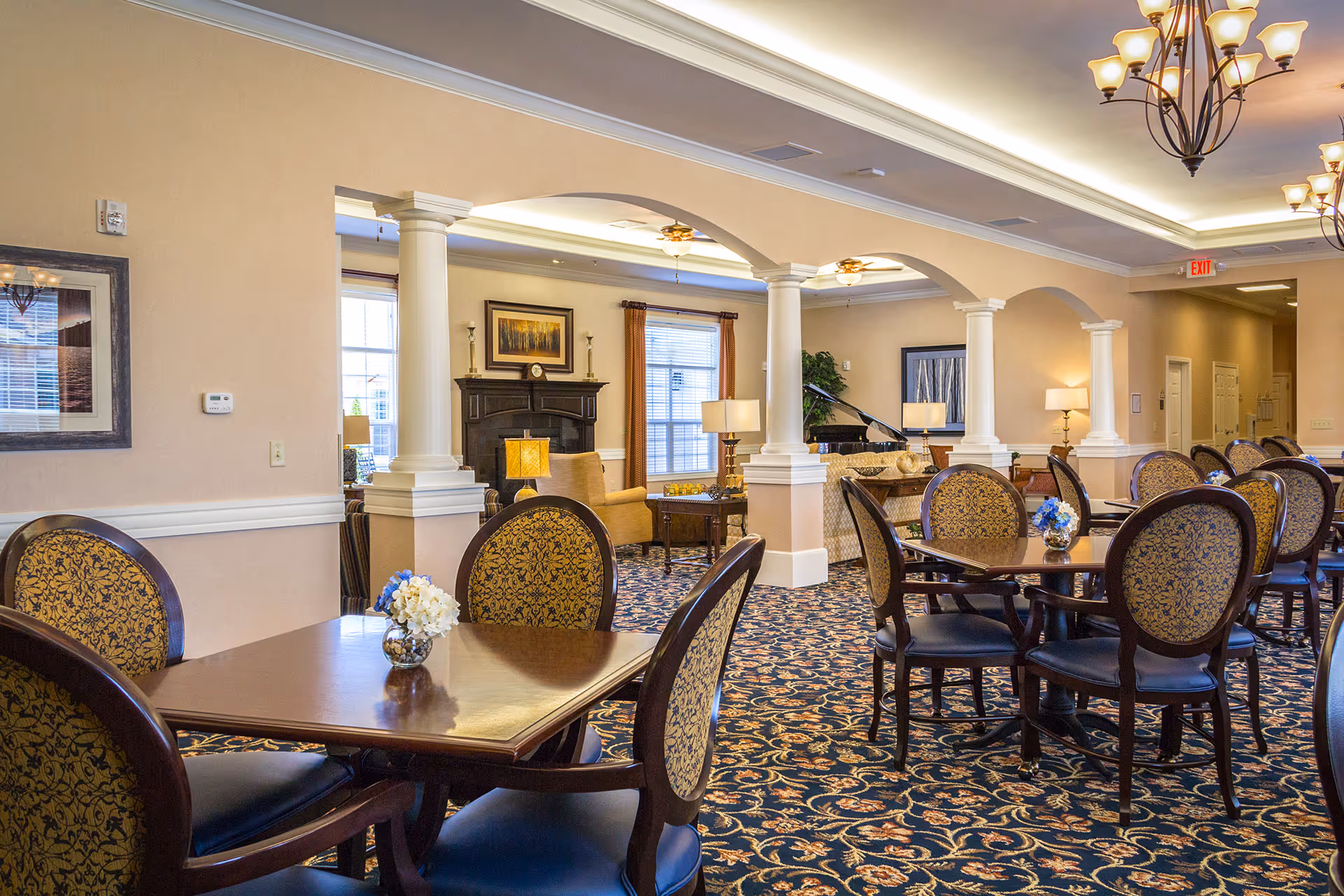A spacious and elegant dining area in Benton House of Blue Springs featuring multiple wooden tables with ornate patterned chairs. The room has a patterned carpet, beige walls, white columns, and warm lighting from chandeliers. In the background, there is a cozy sitting area with armchairs, a fireplace, and a grand piano near windows with curtains.