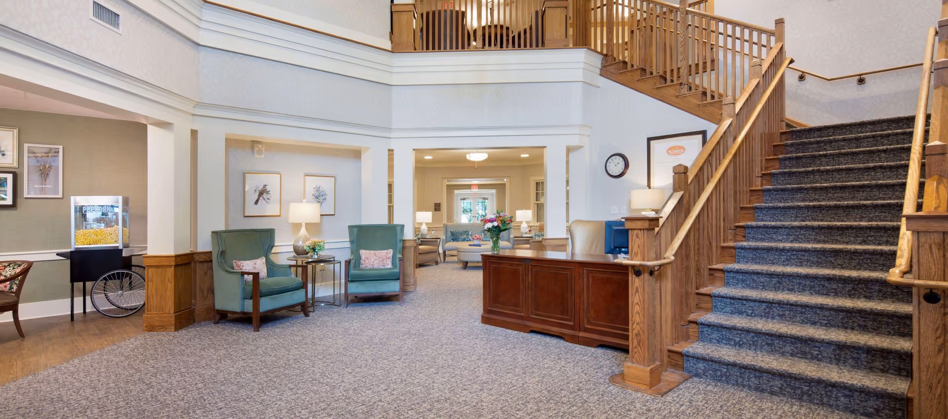 Spacious senior living lobby with a reception desk, seating area, and a carpeted staircase leading to an upper level.