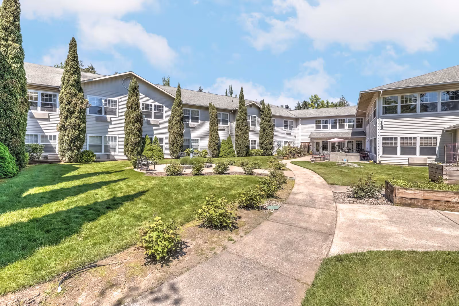 Outdoor courtyard area of Tanner Spring Assisted Living & Memory Care featuring a paved walkway, green lawn, small bushes, tall trees, and a two-story building with many windows under a partly cloudy sky.