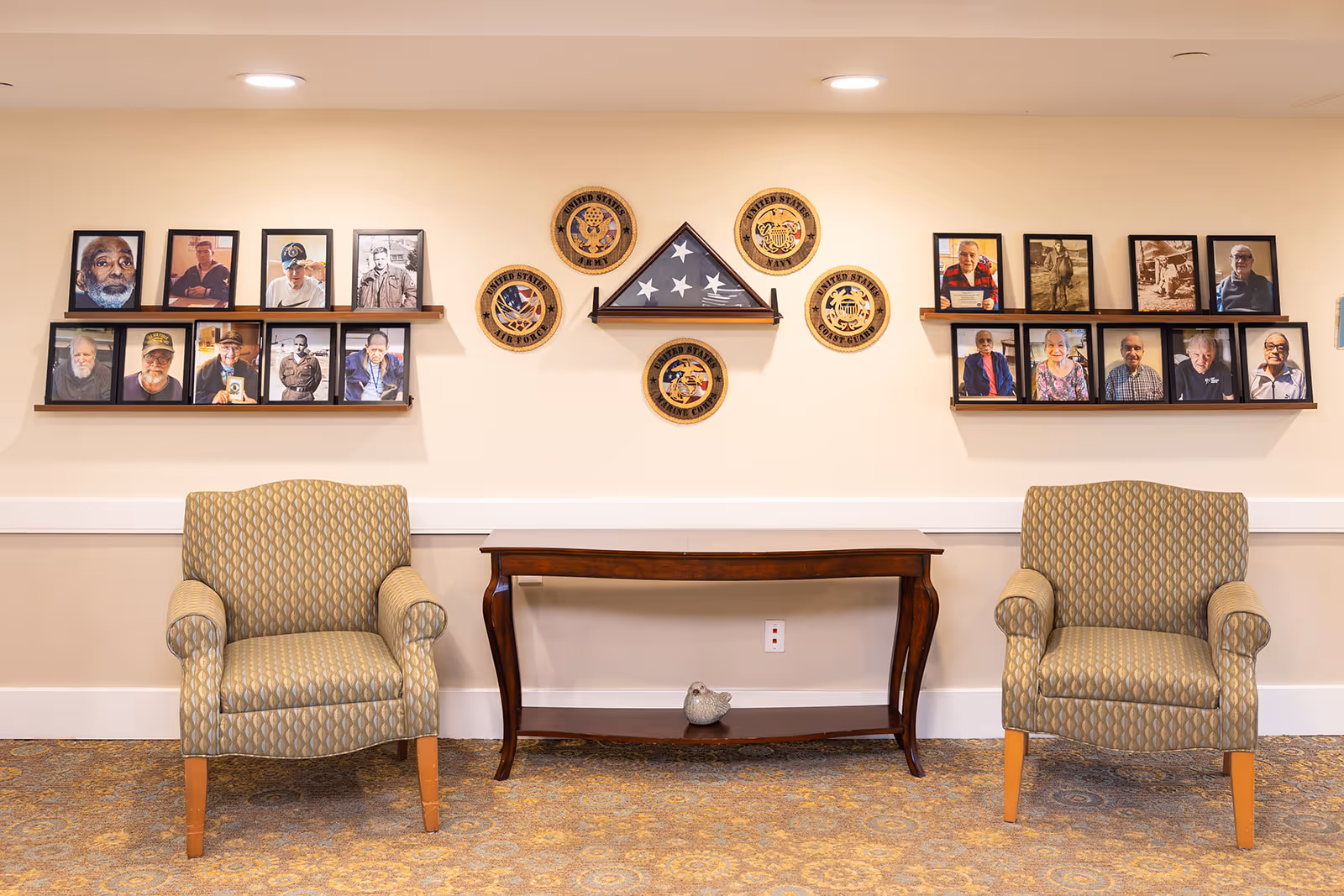 A seating area in a senior living facility with two patterned armchairs on either side of a wooden console table. Above the table on the wall are framed photographs of elderly individuals and circular plaques representing different branches of the United States military, with a folded American flag displayed in the center.