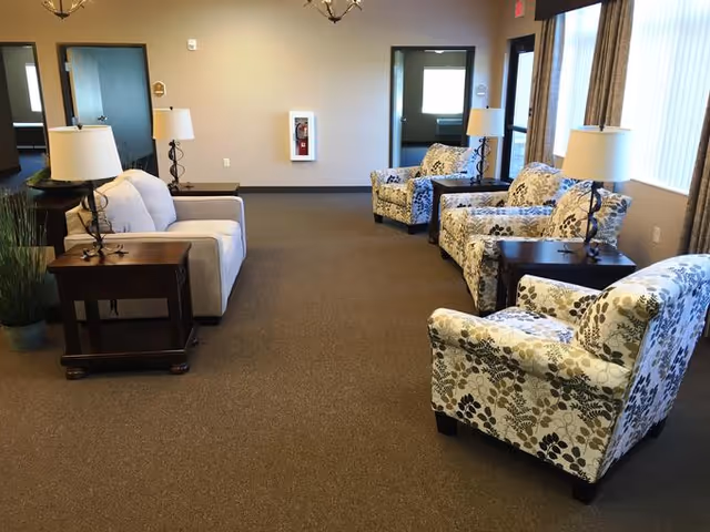 A spacious living room area in an assisted living facility with a beige sofa on the left and three floral-patterned armchairs on the right, each accompanied by a wooden side table with a lamp. The room has beige carpet, cream-colored walls, two doorways in the back, a fire extinguisher mounted on the wall, and large windows with curtains on the right side.