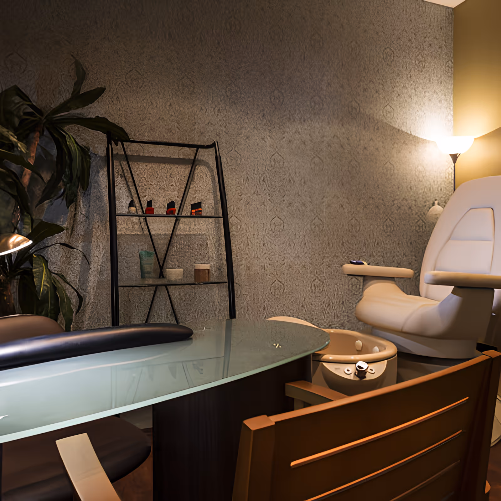 Interior salon area with a glass-topped manicure table, pedicure chair and foot bath beside a shelving unit and lamp.