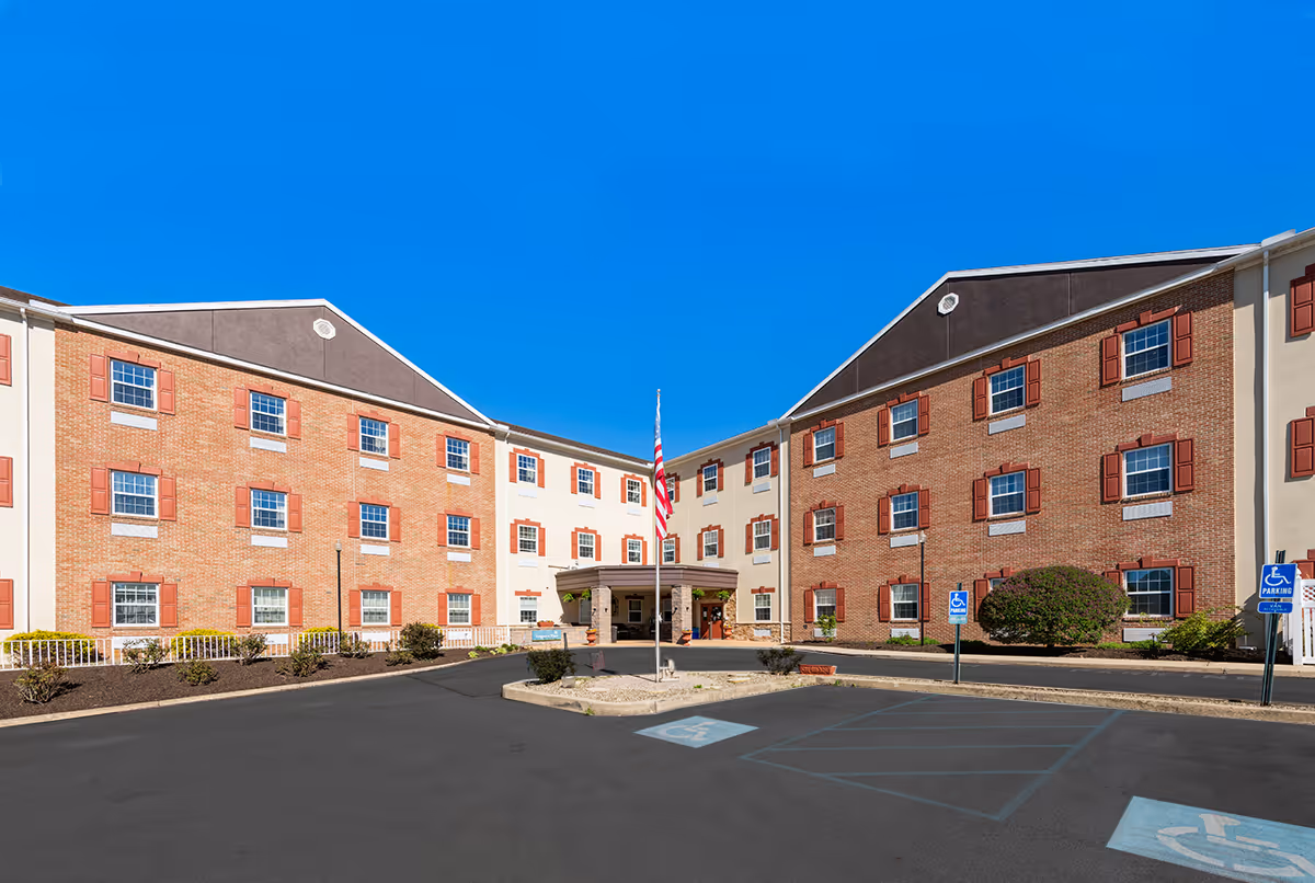 Front exterior view of a three-story senior living facility building with red brick and beige walls, red window shutters, an American flag on a flagpole in the center, and several handicap parking spaces in the foreground under a clear blue sky.