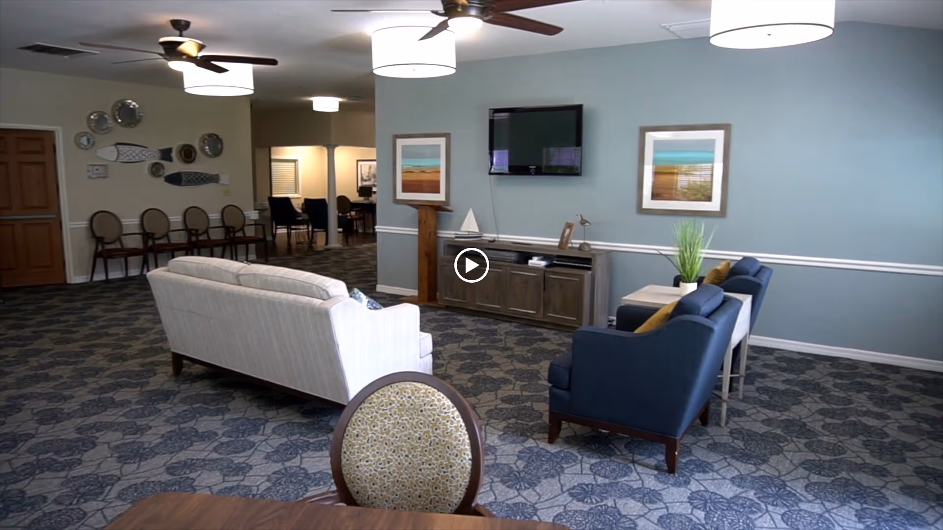 Spacious senior living lounge with sofas and armchairs facing a wall-mounted TV, patterned carpet, and dining chairs along the back.