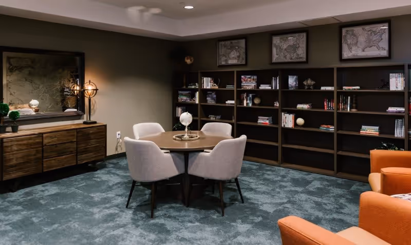 A cozy interior room with a round wooden table surrounded by four upholstered chairs. Behind the table is a large bookshelf filled with books and decorative globes. To the left, there is a wooden sideboard with a lamp and decorative items, and a large framed map on the wall above it. The floor is carpeted in a blue-green color, and two orange armchairs are partially visible on the right side.