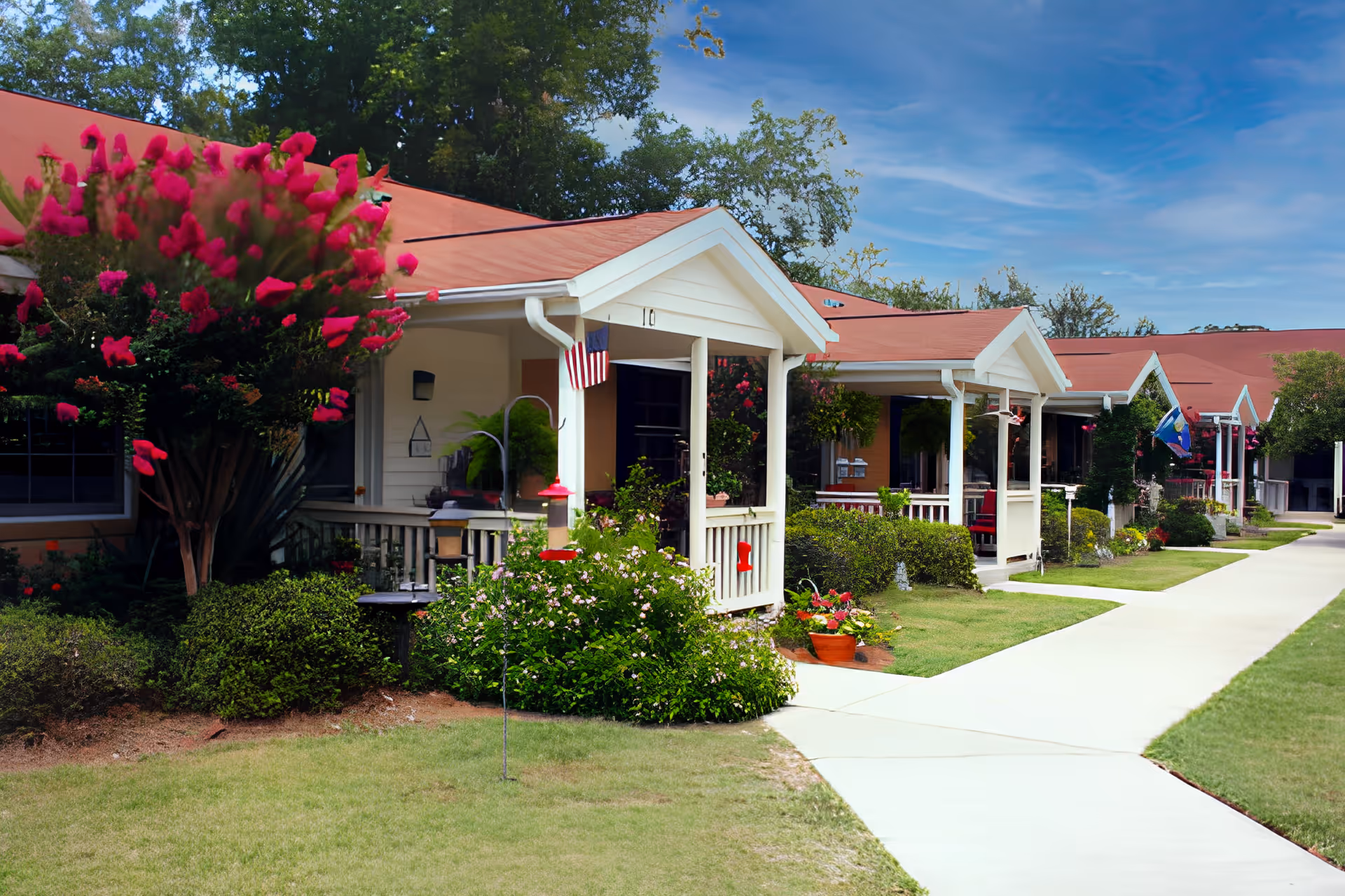 A row of single-story residential units with red roofs and white porches, each decorated with plants and small American flags. A concrete walkway runs alongside the units, bordered by well-maintained grass and shrubs under a blue sky.