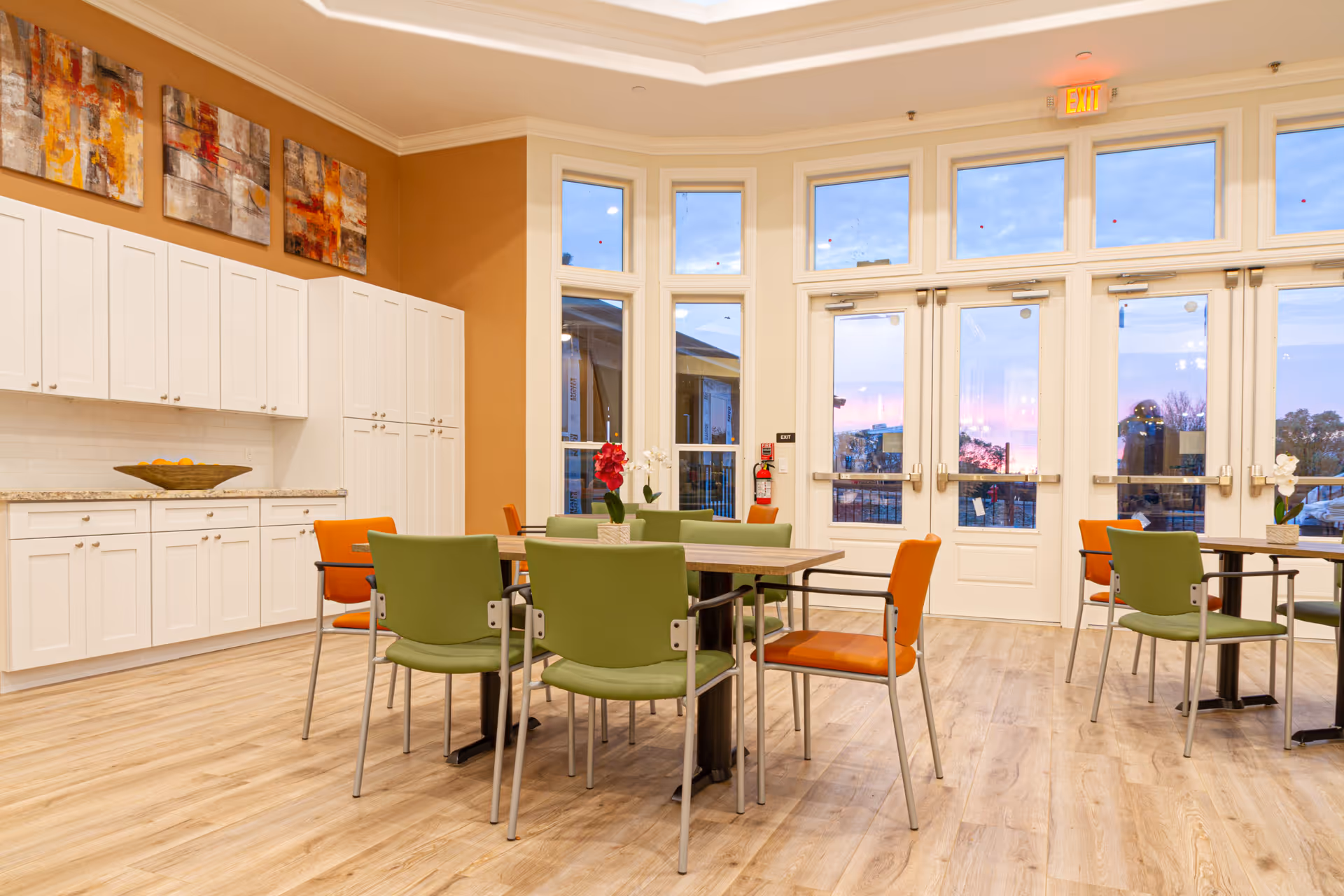 A bright and spacious dining area in a senior living facility with wooden flooring, multiple tables surrounded by green and orange chairs, white cabinets along one wall, and large windows and glass doors letting in natural light during sunset.