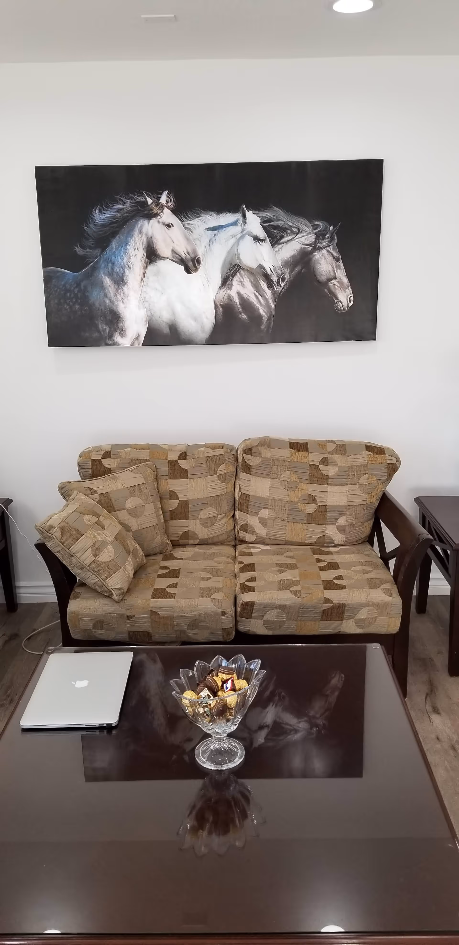 A cozy living room area with a patterned beige sofa adorned with two matching cushions. Above the sofa hangs a large wall art featuring three horses running side by side. In front of the sofa is a dark wooden coffee table with a glass bowl filled with assorted chocolates and a closed silver laptop placed on it. The room has light-colored walls and wooden flooring.