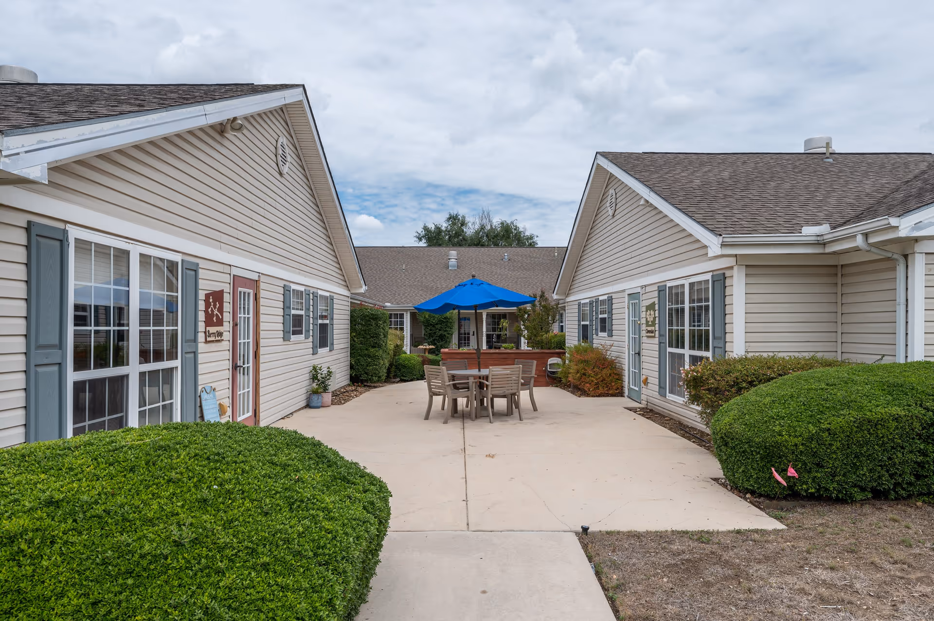 Outdoor courtyard area between two beige buildings with gray shutters. There is a concrete patio with a table and chairs under a blue umbrella, surrounded by green bushes and plants. The sky is partly cloudy.