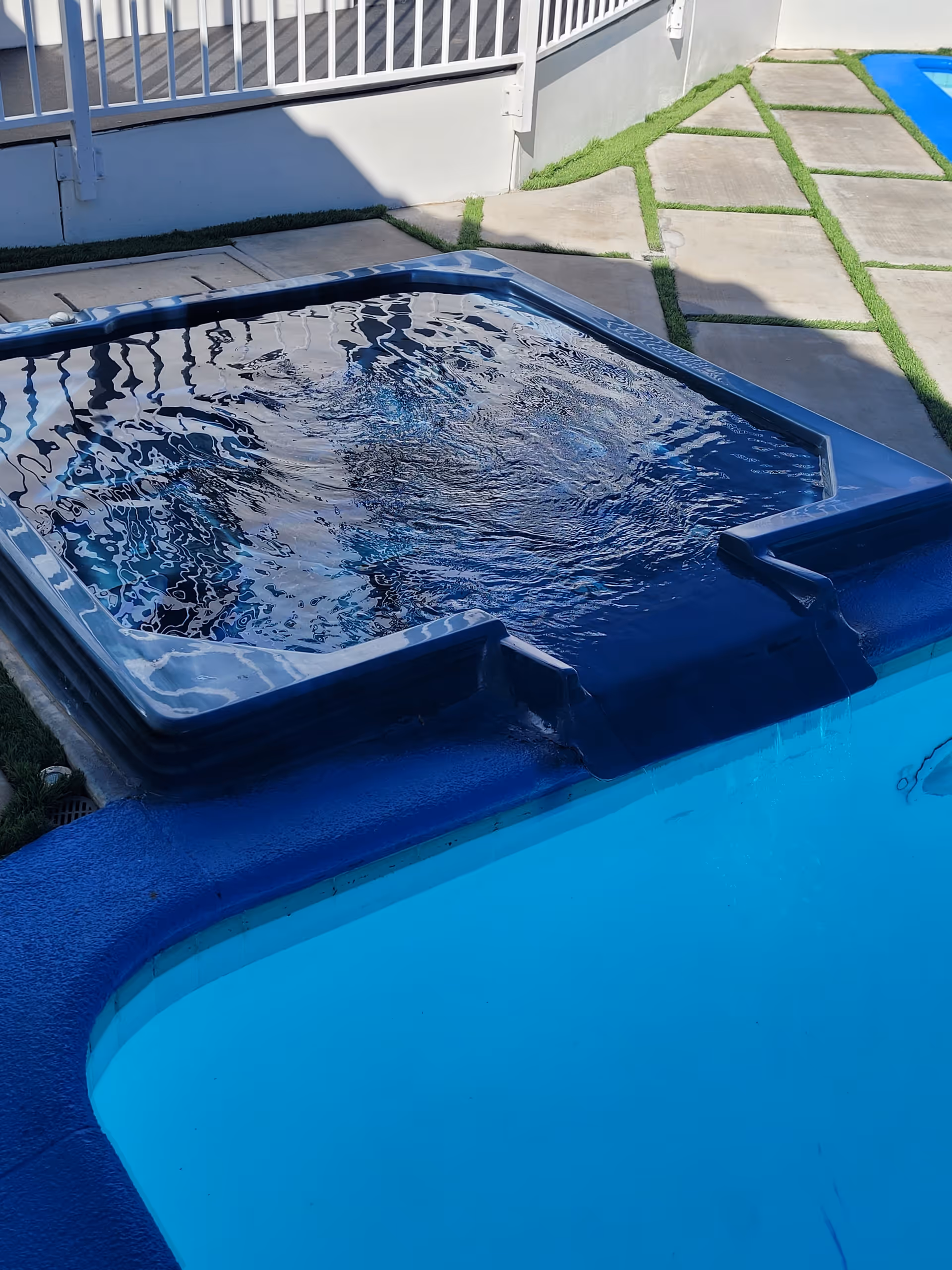 Outdoor area with a blue hot tub adjacent to a swimming pool, surrounded by a tiled patio with grass in between the tiles and a white railing in the background.