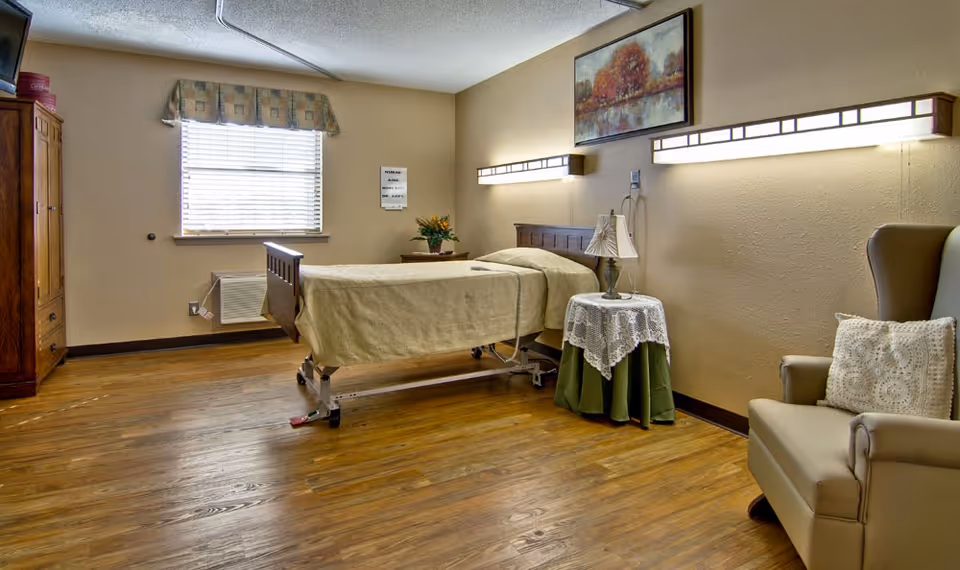 A nursing center bedroom with a single hospital-style bed covered in a beige blanket, a wooden headboard, a small round table with a lace tablecloth and lamp, a cushioned armchair with a decorative pillow, a wooden wardrobe, and a window with blinds and a valance. The room has wooden flooring and beige walls with two long wall-mounted lights and a framed painting above the bed.