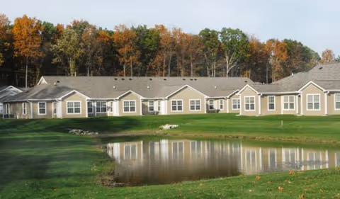 Single-story senior living facility building with beige siding and multiple windows, situated beside a small pond with green grass and trees with autumn foliage in the background.