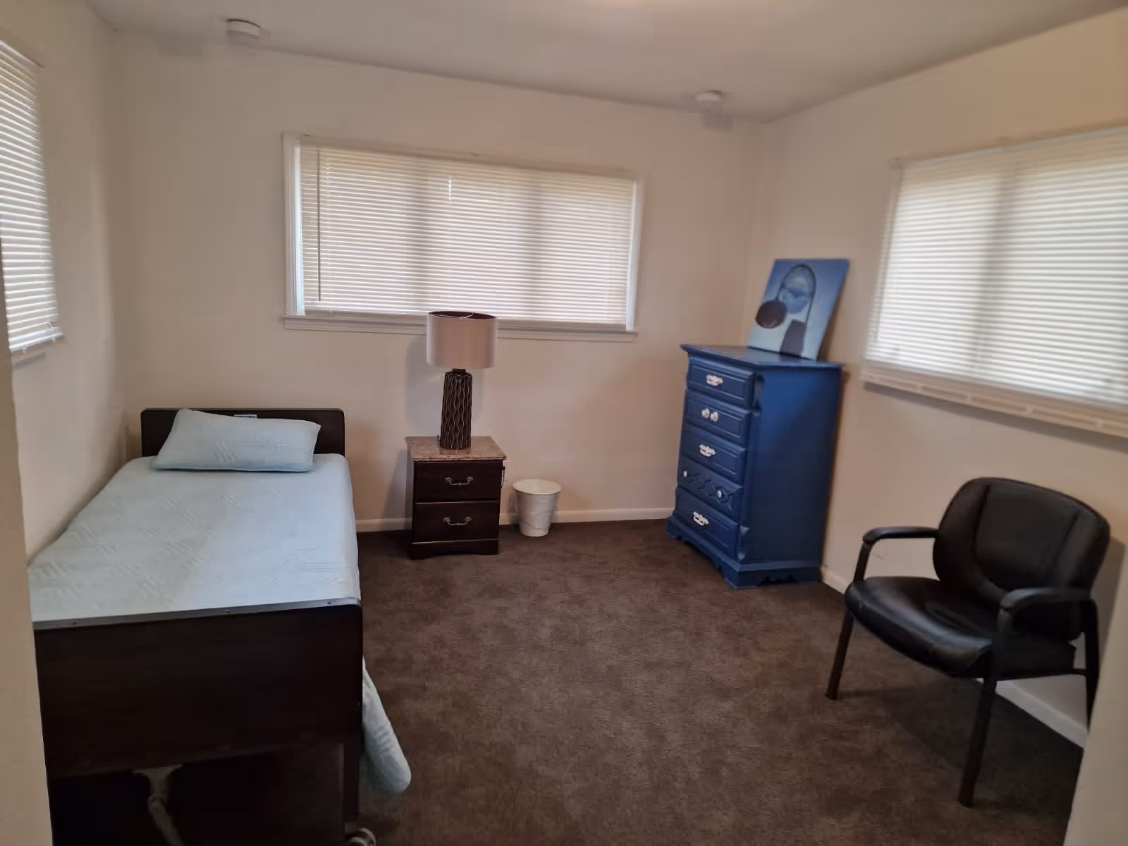 A simple bedroom with a single bed covered with a light blue blanket and pillow, a dark wooden nightstand with a lamp, a blue chest of drawers with a painting on top, a black chair, and two windows with closed blinds.