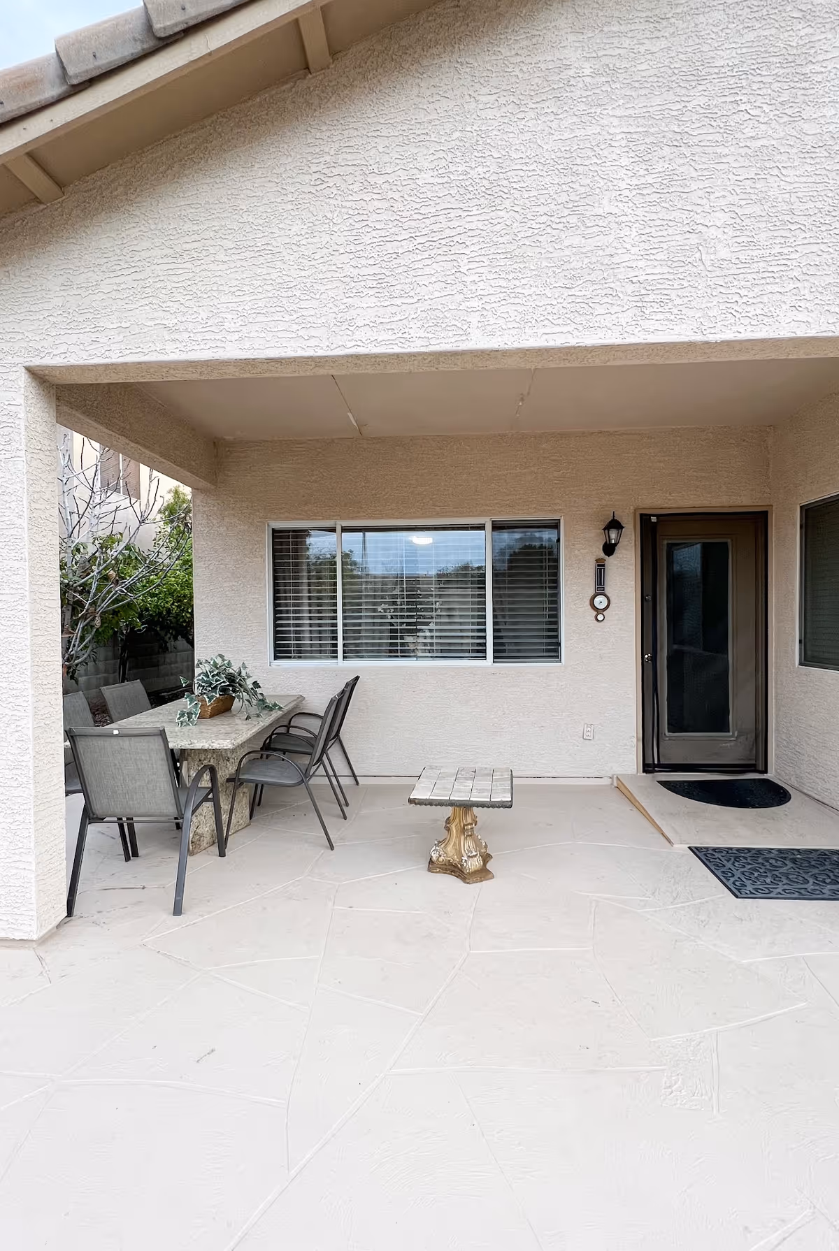 Covered outdoor patio area with a stone table and four chairs, a small decorative table, a window with blinds, a door with a black mat, and a wall-mounted light fixture.