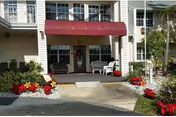 Entrance to a residential building with a red awning above the doorway, white siding, and large windows. There are two white chairs on the porch, red and white flowers planted along the walkway, and a small palm tree to the right.