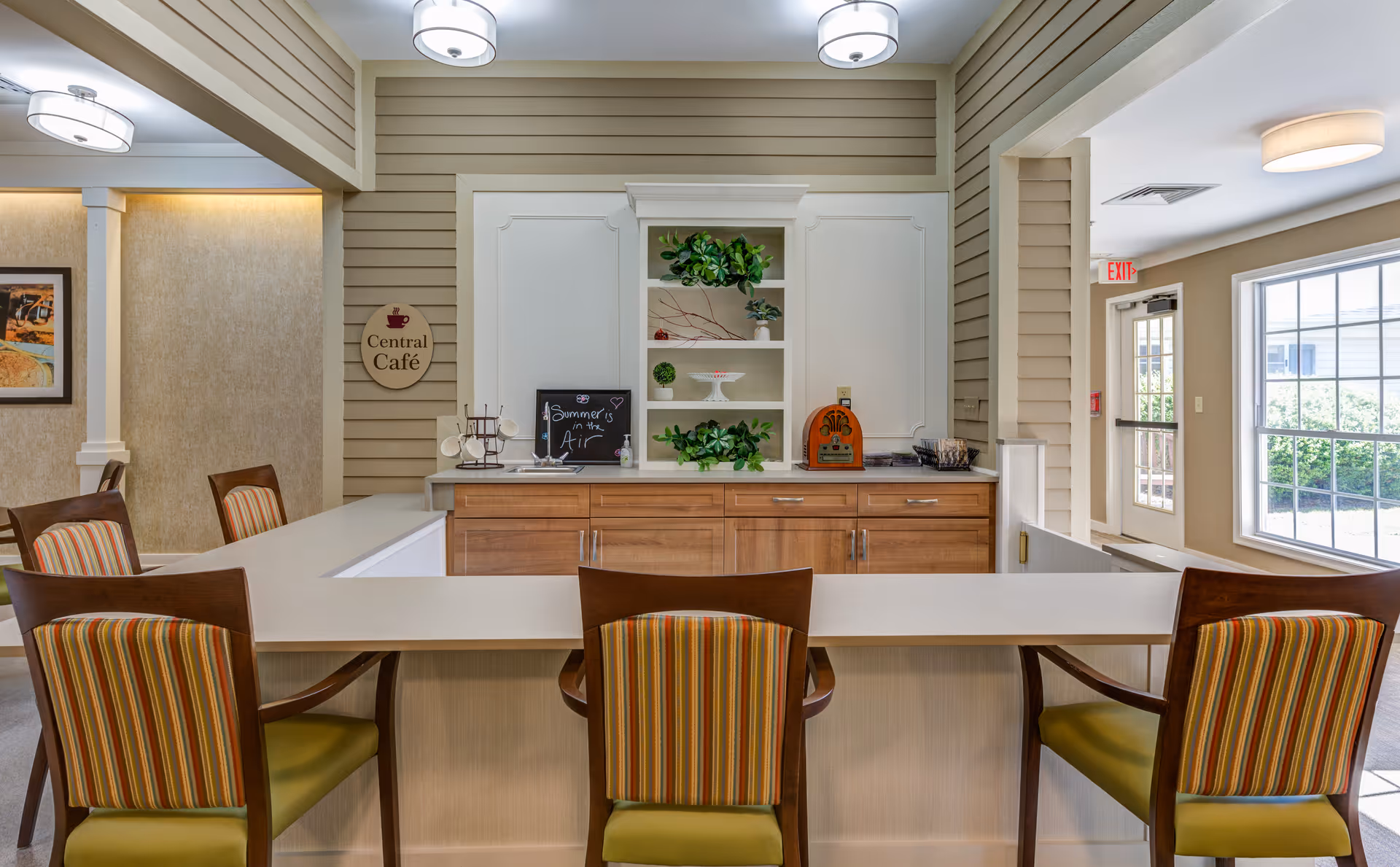 Interior view of a cozy café area with a white countertop and wooden cabinets. Four chairs with striped upholstery are placed around the counter. The back wall features shelves with decorative plants and a small chalkboard sign that reads 'Summer is in the Air'. There is a sign on the wall that says 'Central Café'. Large windows and a door allow natural light to brighten the space.