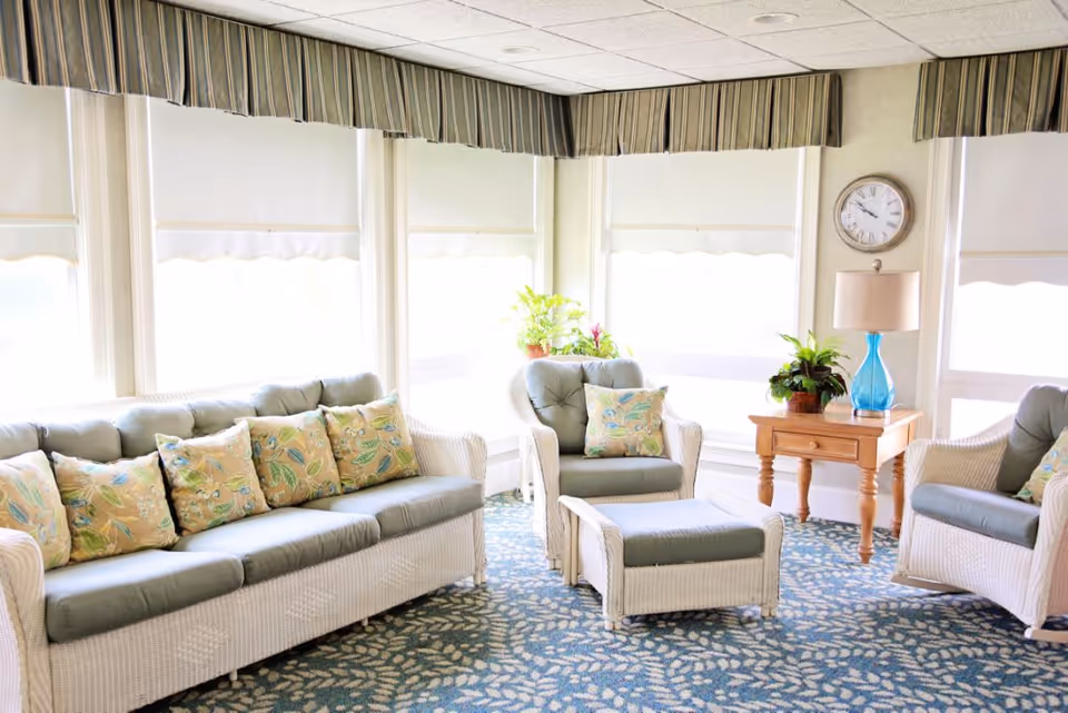 A bright and cozy living room area with white wicker furniture including a sofa, two armchairs, and an ottoman, all with light blue cushions and floral patterned pillows. The room has large windows with white roller shades and striped valances. There is a wooden side table with a blue glass lamp and potted plants, and a wall clock above the table. The floor is covered with a blue patterned carpet.
