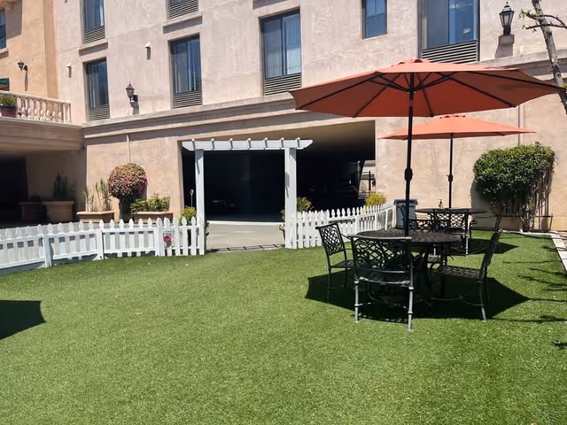 Outdoor courtyard with artificial turf, a white picket fence, metal patio tables and red umbrellas in front of a building facade.