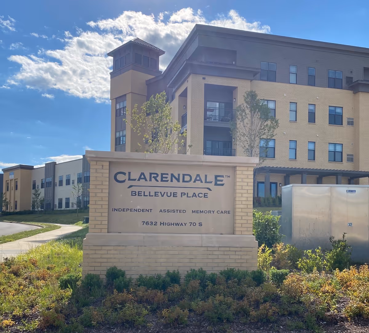 Stone sign reading "Clarendale Bellevue Place" in front of a multi-story senior living building under a blue sky.