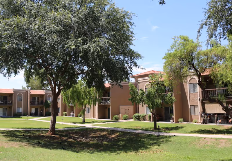Outdoor view of a senior living facility with beige two-story buildings featuring balconies and patios, surrounded by green grass, trees, and walking paths under a clear blue sky.