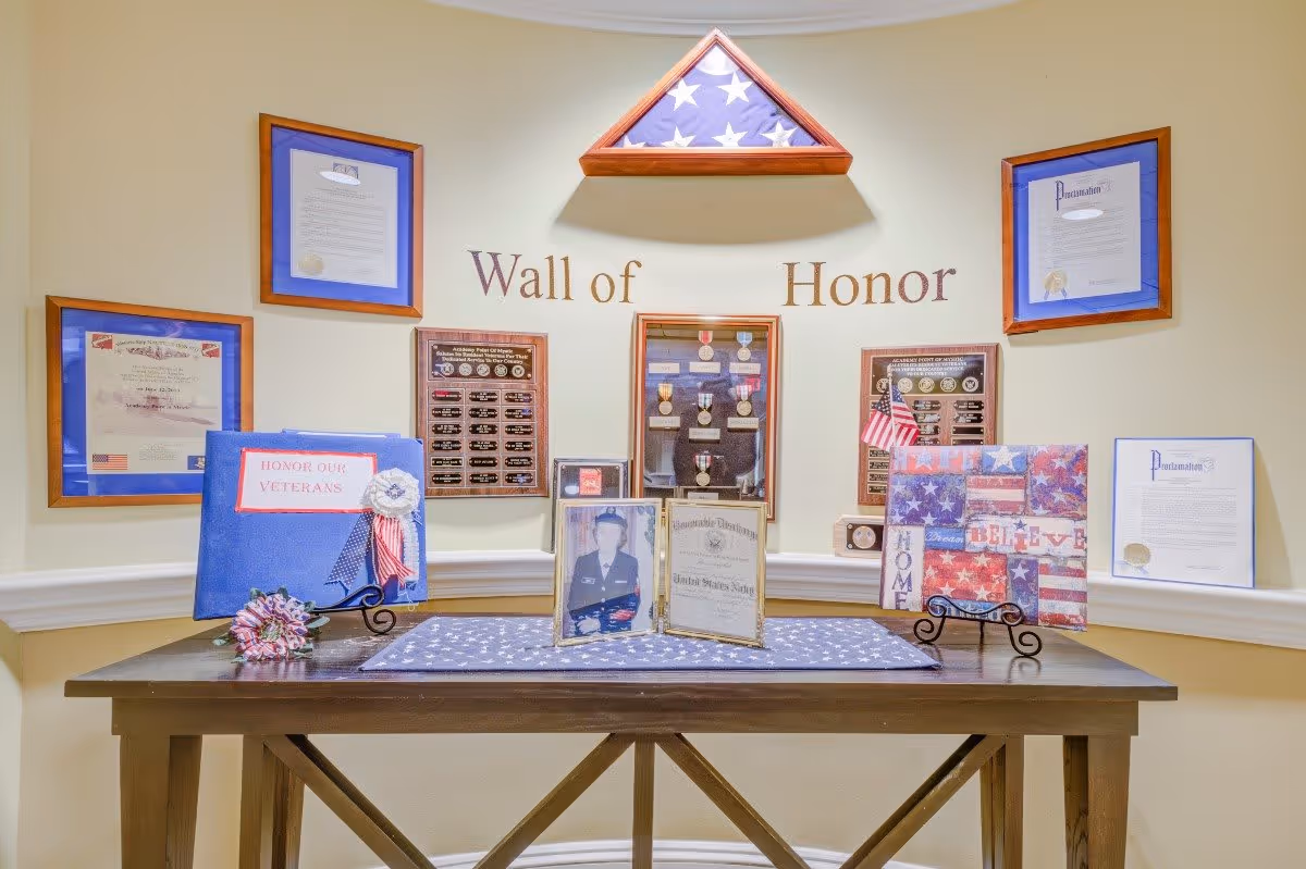 A Wall of Honor display featuring framed certificates, medals, a folded American flag in a triangular wooden case, and patriotic decorations on a wooden table, honoring veterans.