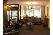 A cozy senior living facility common area with leather sofas and armchairs arranged around a wooden coffee table. The room has large windows letting in natural light, a chandelier hanging from the ceiling, and lamps on side tables. The entrance door is visible on the left side.