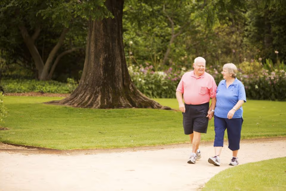 An elderly couple walking hand in hand on a paved path in a lush green park with large trees and flowering bushes in the background.