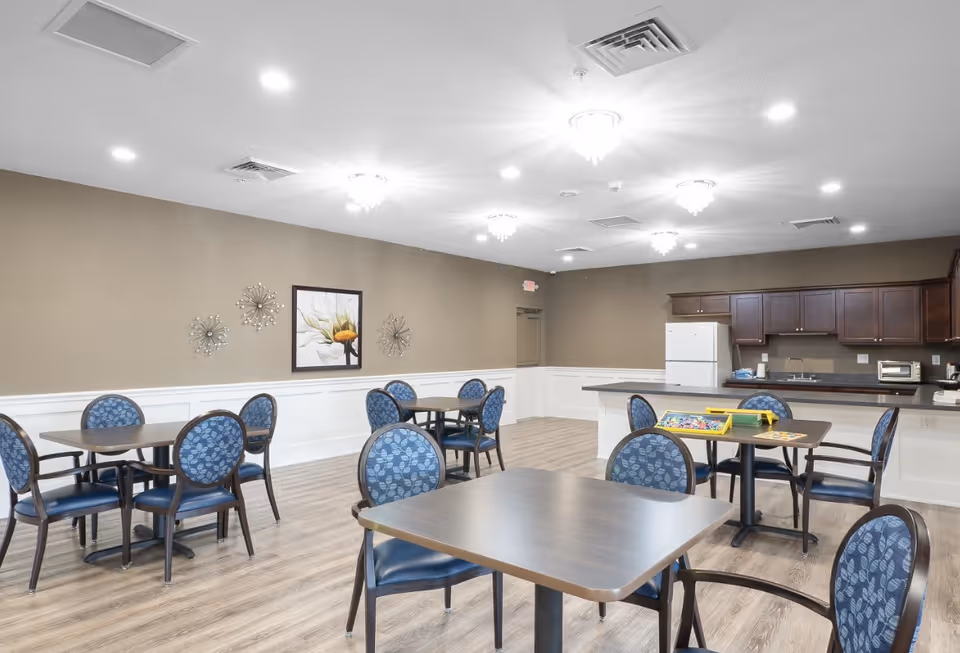 A spacious dining area with several square tables and blue cushioned chairs arranged neatly. The room features wood flooring, beige walls with white wainscoting, and decorative wall art. In the background, there is a kitchen area with dark wood cabinets, a white refrigerator, a microwave, and a sink. The ceiling has multiple recessed lights and small chandeliers providing bright illumination.