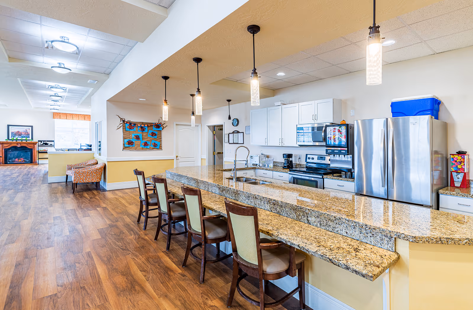 Bright communal kitchen and seating area with a long granite breakfast bar, pendant lights, stainless steel appliances, and wood flooring.