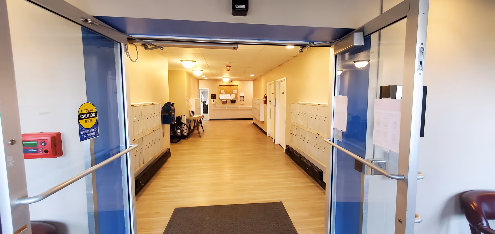 View through open automatic glass doors into a senior apartment lobby hallway with mailboxes along both walls, seating, and a bulletin board at the far end.
