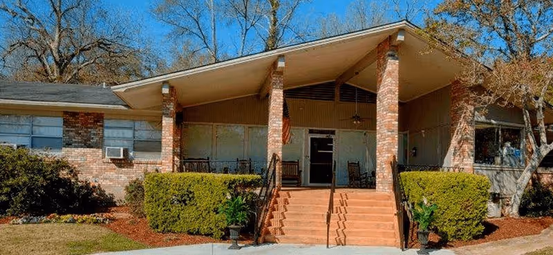 Front exterior view of Natchez Rehabilitation and Healthcare Center showing a single-story brick building with a covered entrance supported by brick columns, stairs leading up to the entrance, and surrounding greenery including bushes and trees under a clear blue sky.