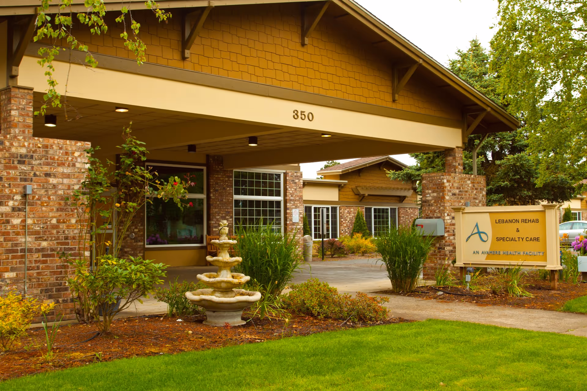 Exterior view of Avamere Rehabilitation of Lebanon building entrance with a covered drop-off area, brick and wood siding, a multi-tiered stone fountain, green landscaping, and a sign that reads 'Lebanon Rehab & Specialty Care, An Avamere Health Facility'.