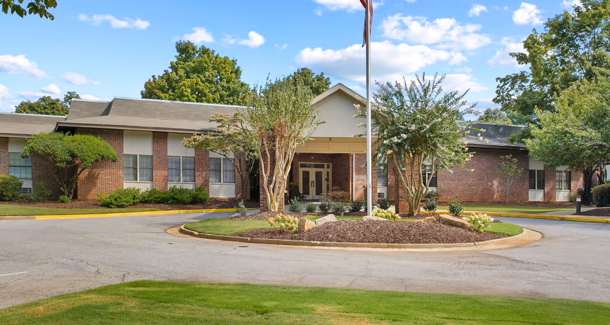 Front exterior view of Mount Vernon Village, a single-story brick building with a circular driveway and landscaped greenery including trees and bushes under a blue sky with scattered clouds.