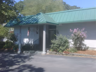 Entrance of a single-story building with a green metal roof, columns, an American flag, and surrounding shrubs and flowers.