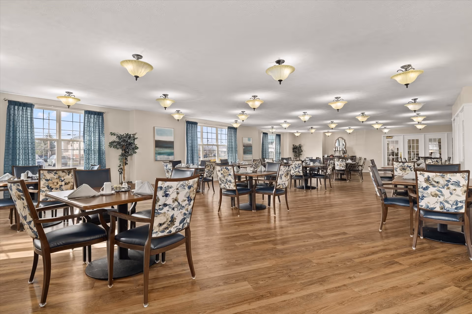 A spacious senior living dining room with multiple wooden tables and chairs featuring floral upholstery. The room has large windows with blue curtains allowing natural light to fill the space. The ceiling is adorned with multiple light fixtures, and the floor is covered with wood-style flooring. There are paintings and plants decorating the walls and corners.