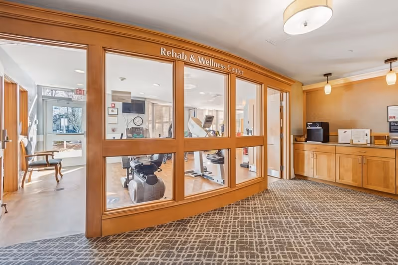 Interior view of a rehab and wellness center with exercise equipment visible through large glass windows framed in wood. The room outside the center has patterned carpet flooring, wooden cabinets with a coffee machine, and pendant lighting. A hallway with chairs and a door leading outside is visible to the left.