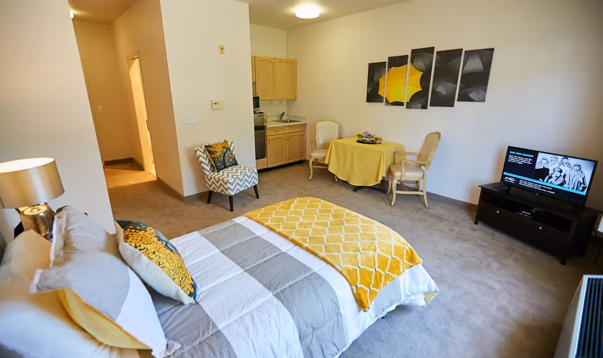 A bright studio apartment with a bed in the foreground, a small kitchenette and dining table, armchairs, and a TV.