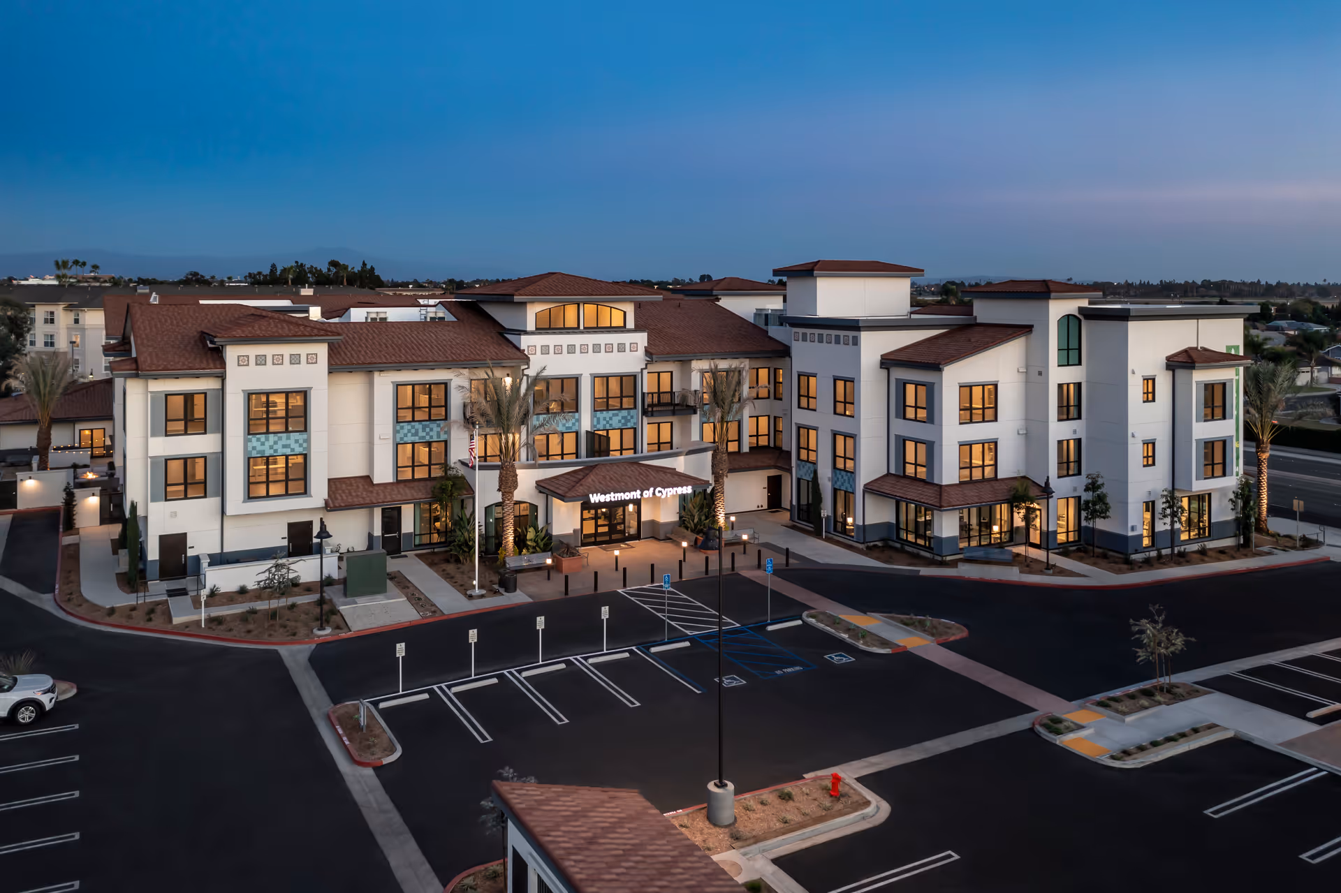 Front exterior of a three-story senior living building with an illuminated entrance and mostly empty parking lot at dusk.