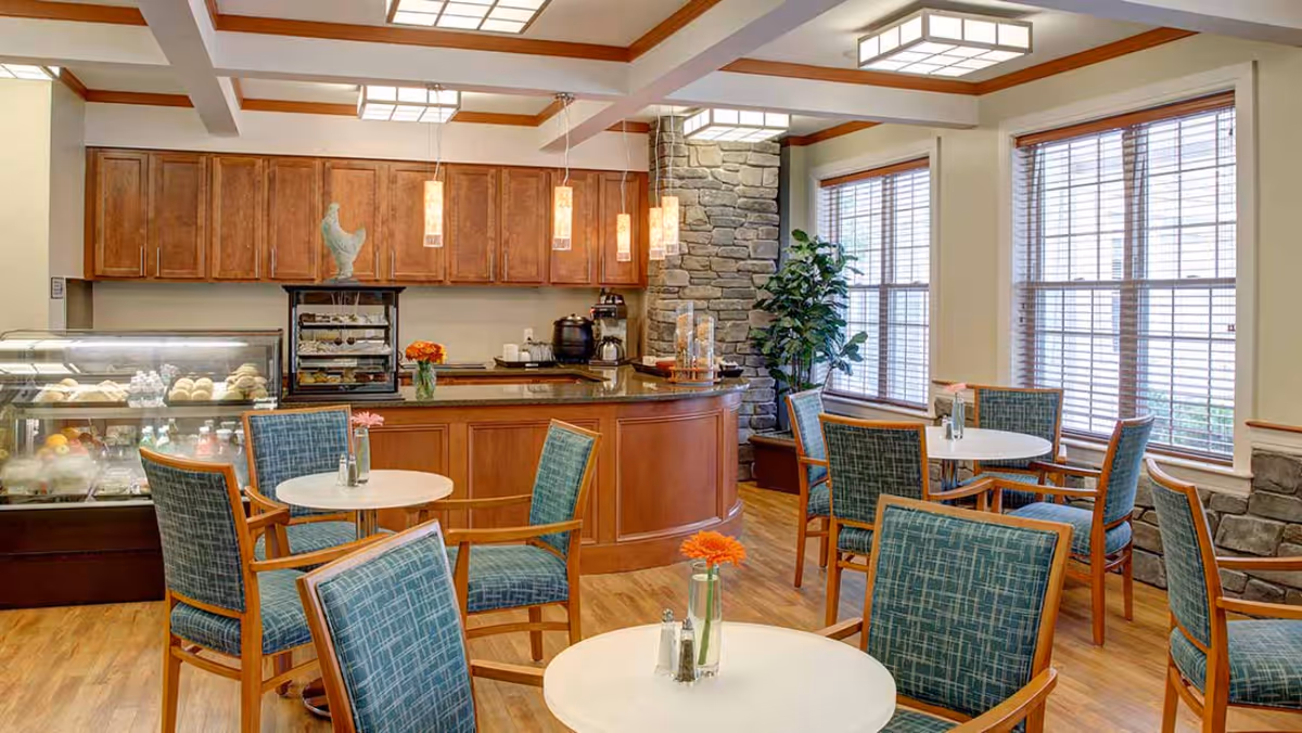 A cozy dining area in a senior living facility featuring round tables with blue upholstered chairs, small flower vases on each table, large windows with wooden blinds, a stone accent wall, and a counter area with wooden cabinets and a display case containing baked goods.
